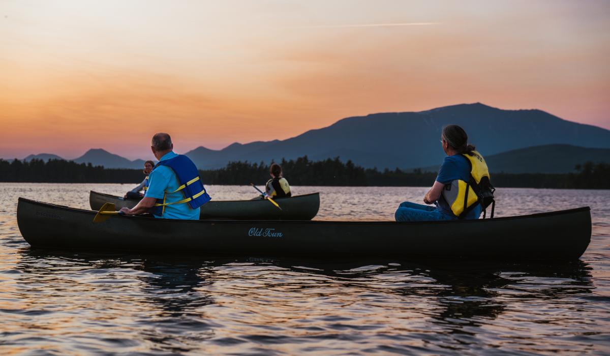 Native and nonnative people traveling in two canoes with backs to the camera and sunset over mountains in the distance.