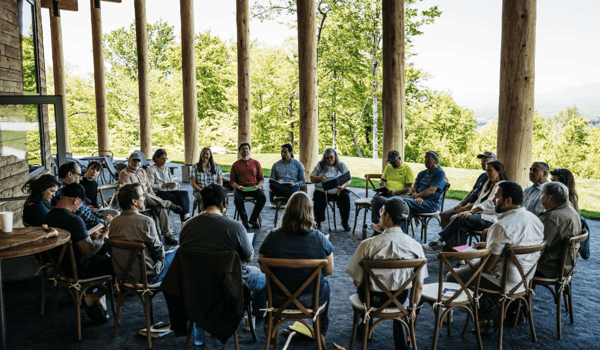 22 Wabanaki and Non-native leaders sit together in discussion. they are inside a spacious room with a view of a bright landscape showing trees in the foreground with Katahdin and other mountains in the distance.