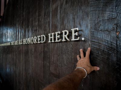 Hand in front of text on a wall saying "We are all honored here"