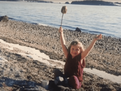 Ann Ranco, as a small girl in cozy red vest and pants, sits on the calm shore at Redman's Beach waving a giant toasted marshmallow above her head and grinning at her family who are behind the camera.