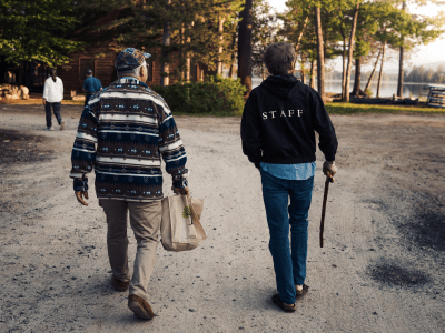 A Wabanaki elder and a non-Native elder walk together on a path alongside trees and a lake on a cool morning in early summer.