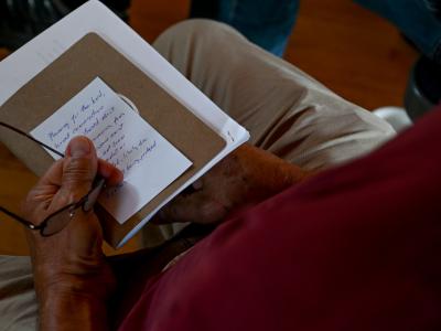 a pair of hands holding glasses, a notebook, and an index card with thoughts and commitments from a day of discussion.