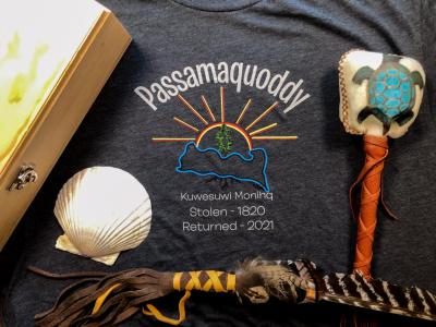 Several ceremonial items like a box, a shell, and a feather rest on a tshirt that says, "Passamaquoddy. Kuwesuwi Monihq. Stolen 1820. Returned 2021." and shows an embroidered image of a pine tree, surrounded by water, standing in front of the sun.