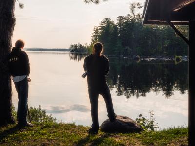 two tall people standing on the bank of a perfectly calm lake with trees in the distance in the early morning, sharing a moment of reflection. They are facing away from the camera and are both looking at something along the shore, one leans agains a large pine trunk.