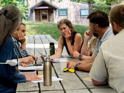 a group of six people gather sitting at a picnic table with pens, notebooks, and travel mugs, thoughtfully discussing and sharing ideas with each other on a warm sunny day.
