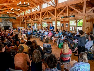 a view from 15 feet off the ground of a large group of conservation community leaders gathered in a large room with a high ceiling. They are taking turns sharing their experience of being involved with land return efforts in solidarity with Wabanaki.