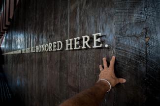Hand in front of text on a wall saying "We are all honored here"