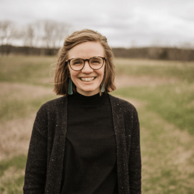 a young pale-skinned woman wearing a dark sweater and glasses stands smiling in a pasture on a cold cloudy day.