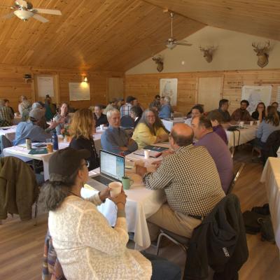 groups of people, both Wabanaki and non-Wabanaki, sit at tables with notebooks and papers discussing a wide variety of issues.